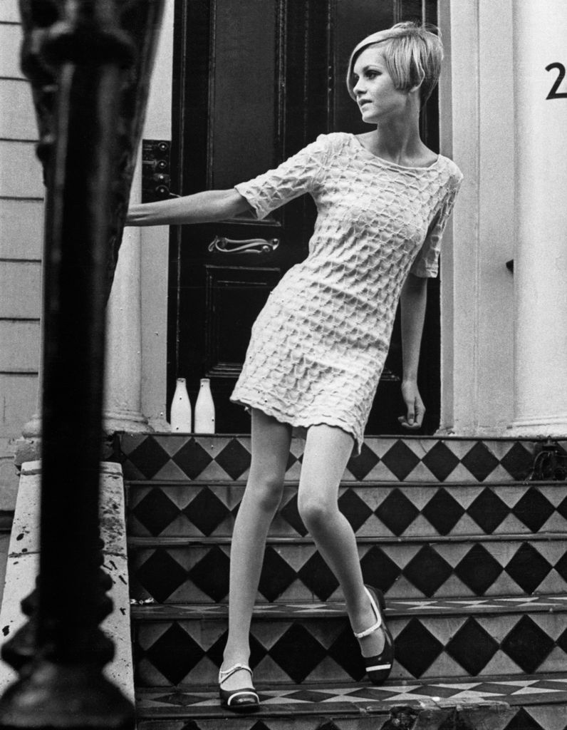 Twiggy steps confidently on a checkered staircase in a white textured mini dress. The monochrome scene highlights her modern silhouette and minimalist styling