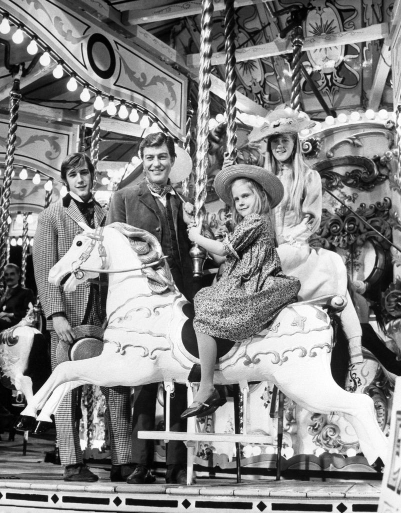 Six-year-old CARRIE BETH and her sister STANCEY, 13, steal the scene here from their brother BARRY, 16, and father DICK VAN DYKE, who watch as the girls go round the merry-go-round at Pinewood Studios. Three of the star's children thus got into one of the