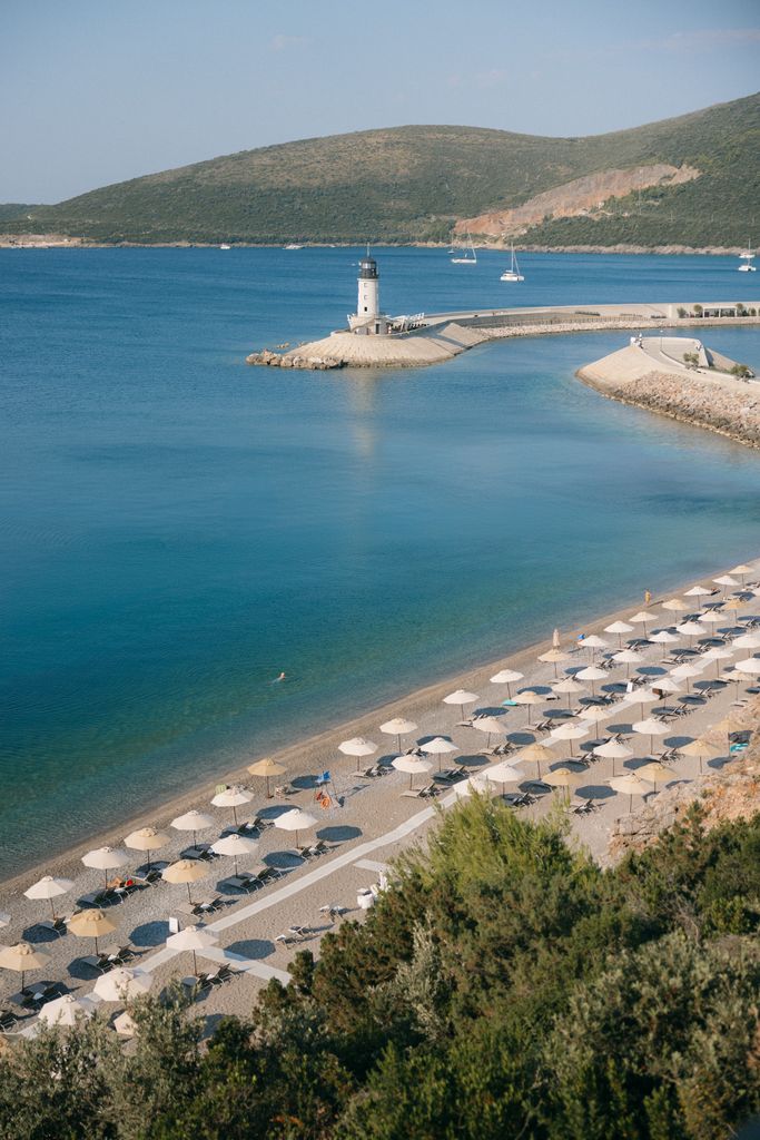 Lustica Bay, Monténégro, propose une plage avec des parapluies et des chair de poule, un phare blanc sur une jetée en pierre et une eau bleue calme avec des bateaux lointains, tous contre des collines vertes.