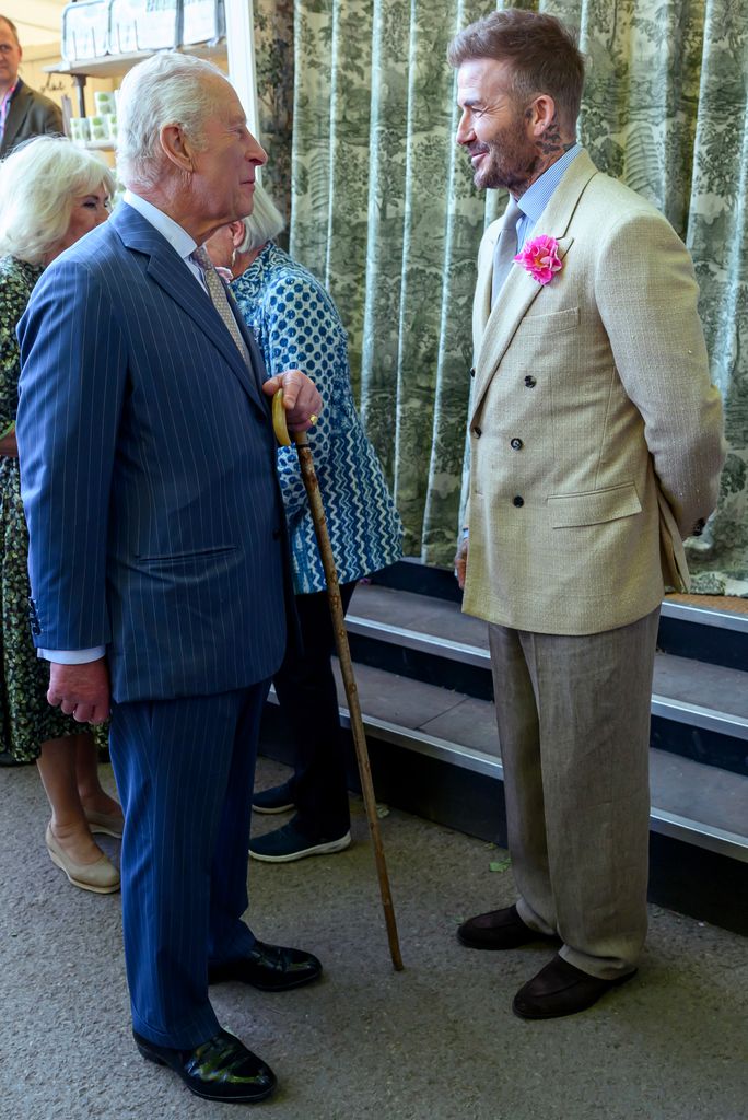 David Beckham wearing a beige double-breasted suit with a pink flower and blue shirt, standing beside King Charles in pinstripes at the Chelsea Flower Show 2025, both stylishly dressed.