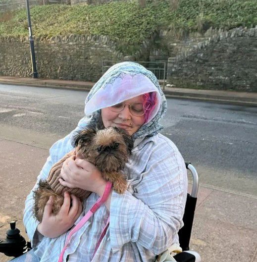 betty ross holding pet dog wearing a raincoat and sitting in wheelchair by side of road