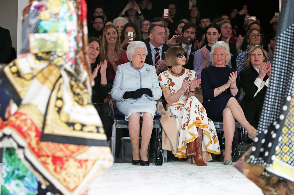The Queen looking surprised next to Anna Wintour and Angela Kelly as they are sat watching a fashion show