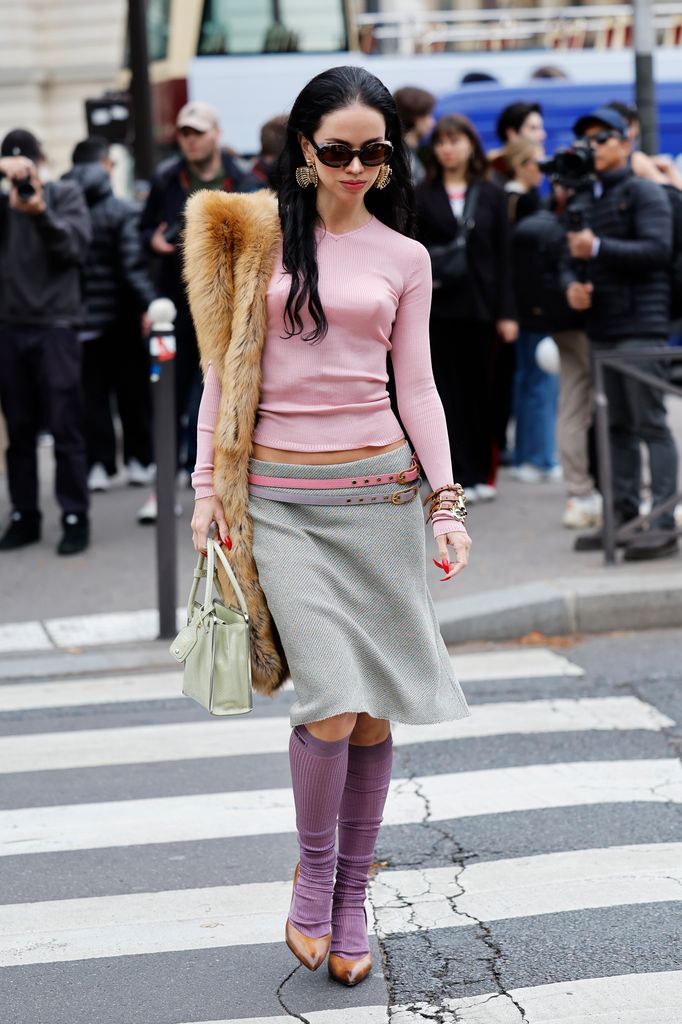 A guest wears pink long sleeves top, grey midi skirt with pink and lilac leather belts, purple socks, brown heels, green bag, beige fur scarf, outside Miu Miu, during the Womenswear Spring Summer 2026 as part of Paris Fashion Week