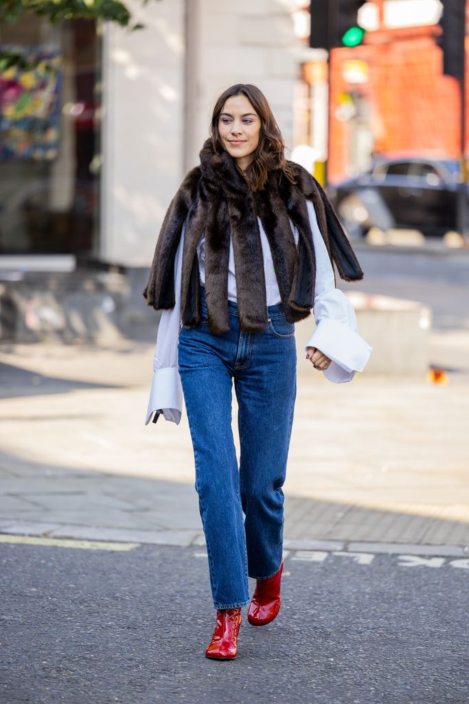 Alexa Chung wears fur cape with slits, denim jeans, white button shirt, red ankle boots outside JW Anderson during London Fashion Week September 2023 at the on September 16, 2023 i
