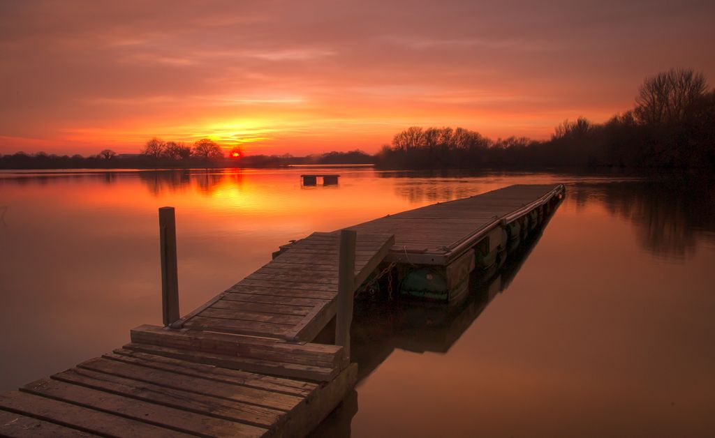 Picture of a floating wooden pier with the sunset across the lake in the background. Black swan lake - Dinton Pastures - Wokingham
