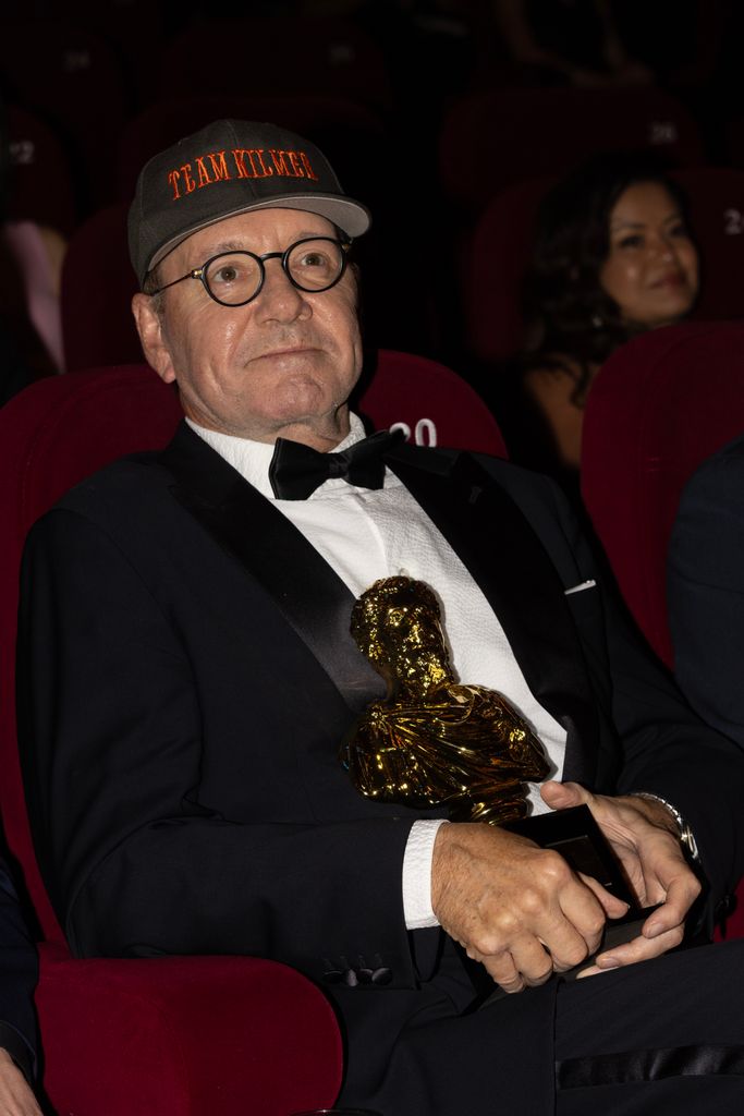 Kevin Spacey holding an award wearing a baseball hat looking stern