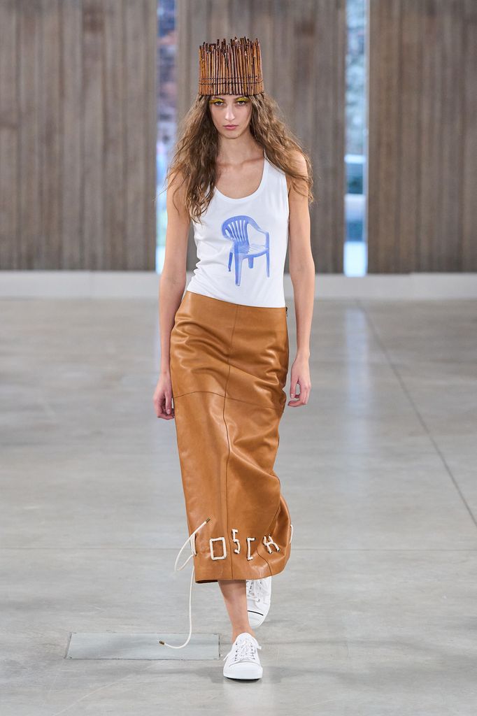 A model wears a tan leather pencil skirt and white vest. The skirt has Moschino crudely stitched into the hem