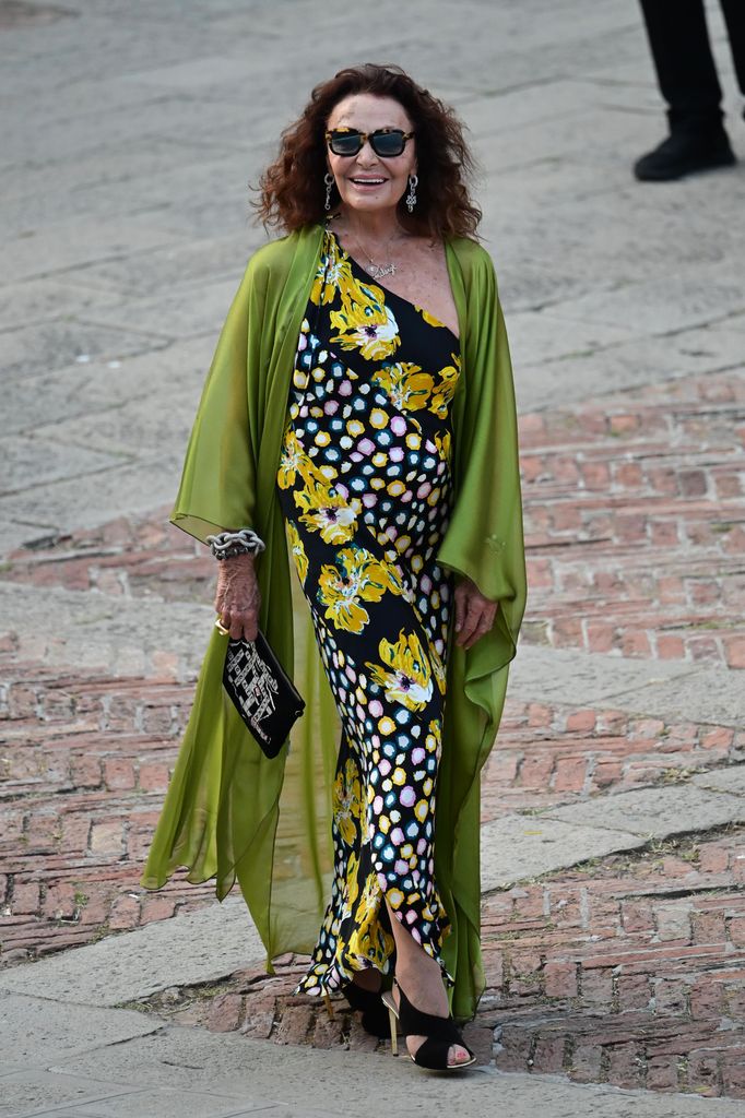 Diane von Furstenberg wears a one-shoulder dress with floral and polka-dot patterns, layered with a sheer green shawl and accessorised with sunglasses and a statement bracelet.