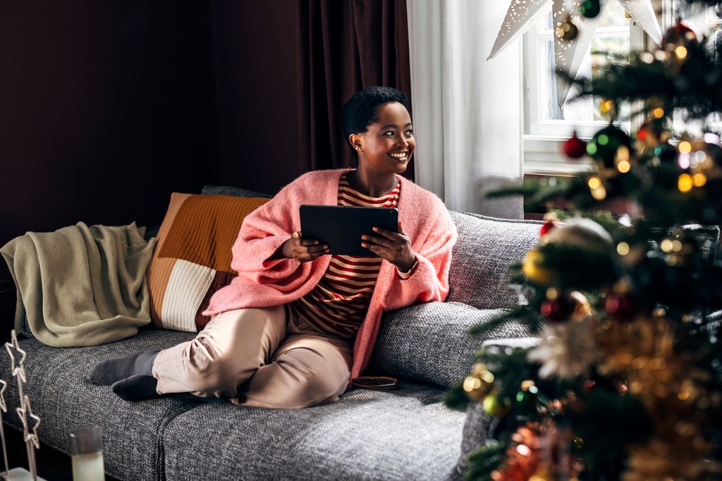  woman comfortably sitting on the sofa in her living room, enjoying Christmas holidays, spending her time browsing social media on a tablet device.