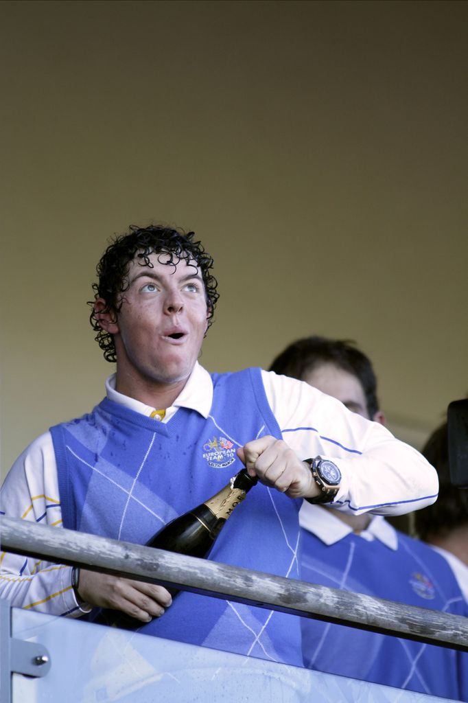  Rory McIlroy popping open a bottle of champagne wearing a purple sweater vest layered over a crisp white polo shirt.
