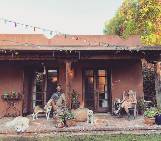 a man and woman sit on separate chairs on the red brick patio of a Spanish style bungalow decorated with colourful lights in the soft sunshine as dogs rest on the ground closeby