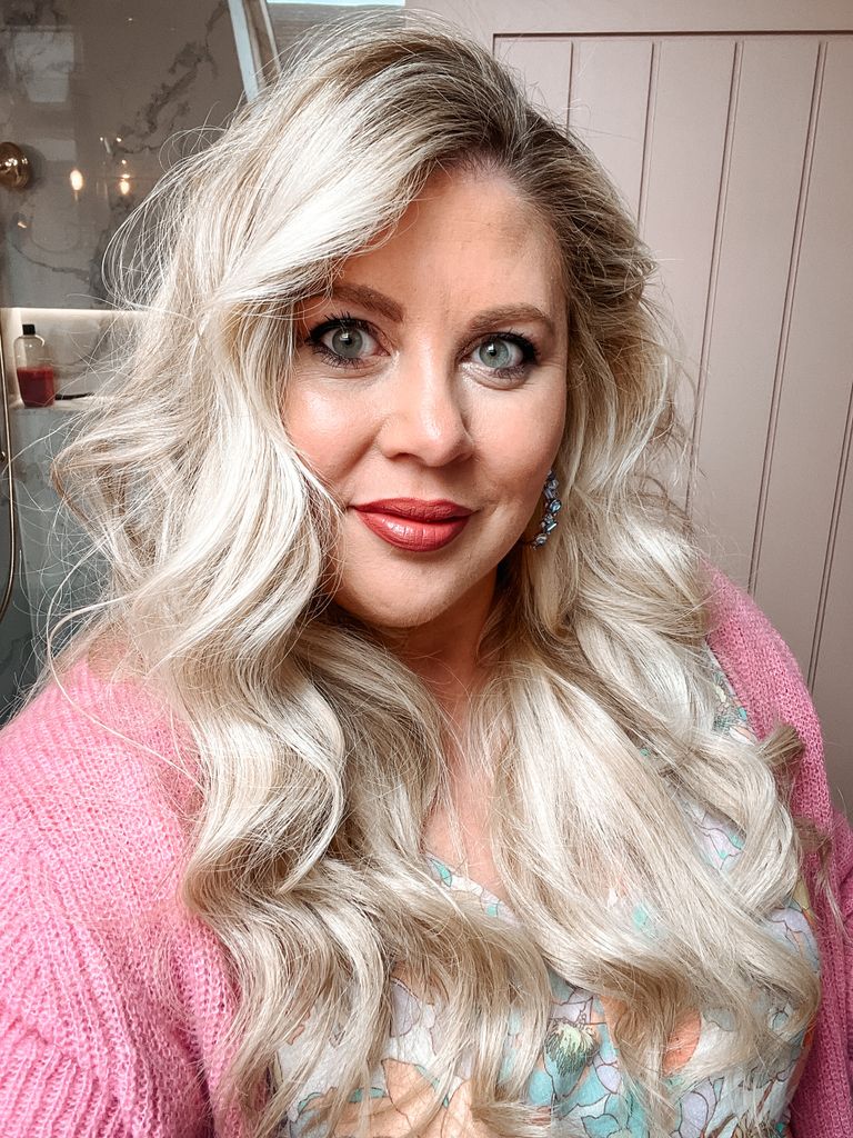 selfie of a woman with blonde hair wearing a pink cardigan