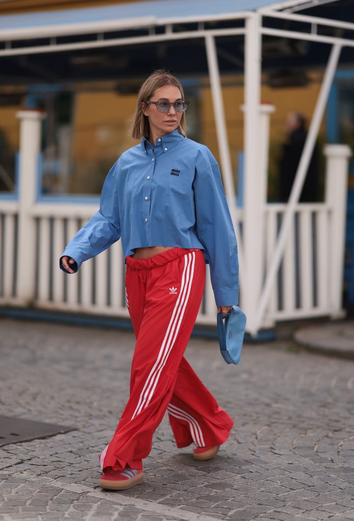 Karin Teigl seen wearing Rhude grey sunglasses, Miu Miu blue cotton cropped buttoned shirt, Adidas red long wide leg track pants, Prada Re-edition blue nylon bag and Adidas Gazelle red / blue suede leather platform sneakers, o