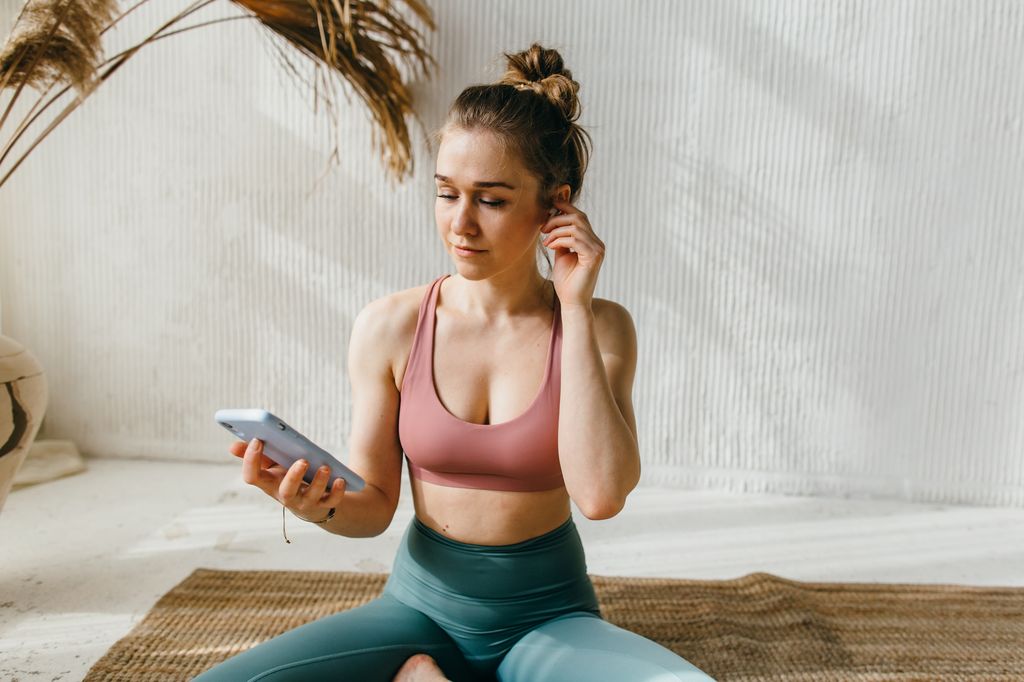 Young woman wearing wireless headphones uses her smartphone to set up a meditation app, sets up a music playlist before a workout, and selects a suitable fitness program online