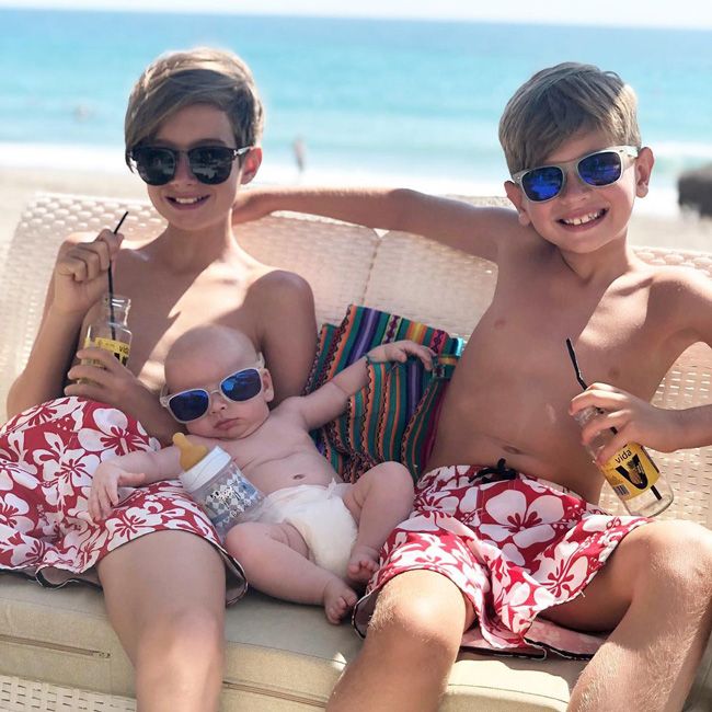 two young boys sit on a beach chair in the sunshine wearing sunglasses with a baby positioned between them wearing a nappy and matching sunglasses