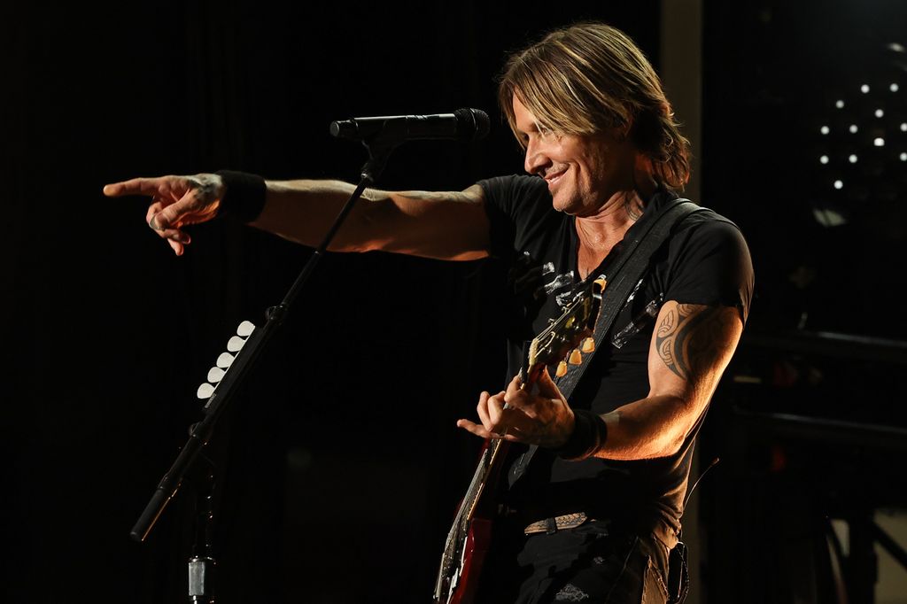 Keith Urban facing left performing on stage with a guitar and pointing out at the crowd