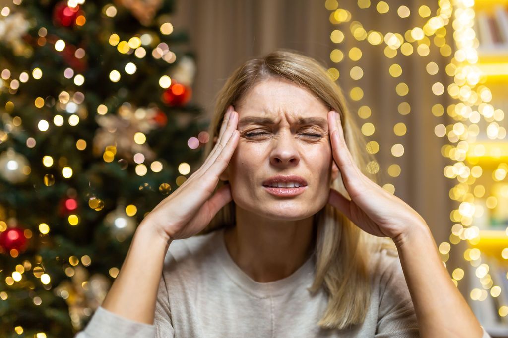 A young beautiful woman is sitting at home, holding her head with her hands, grimacing in pain. She feels a severe headache. She is tired of preparing for the New Year and Christmas holidays.