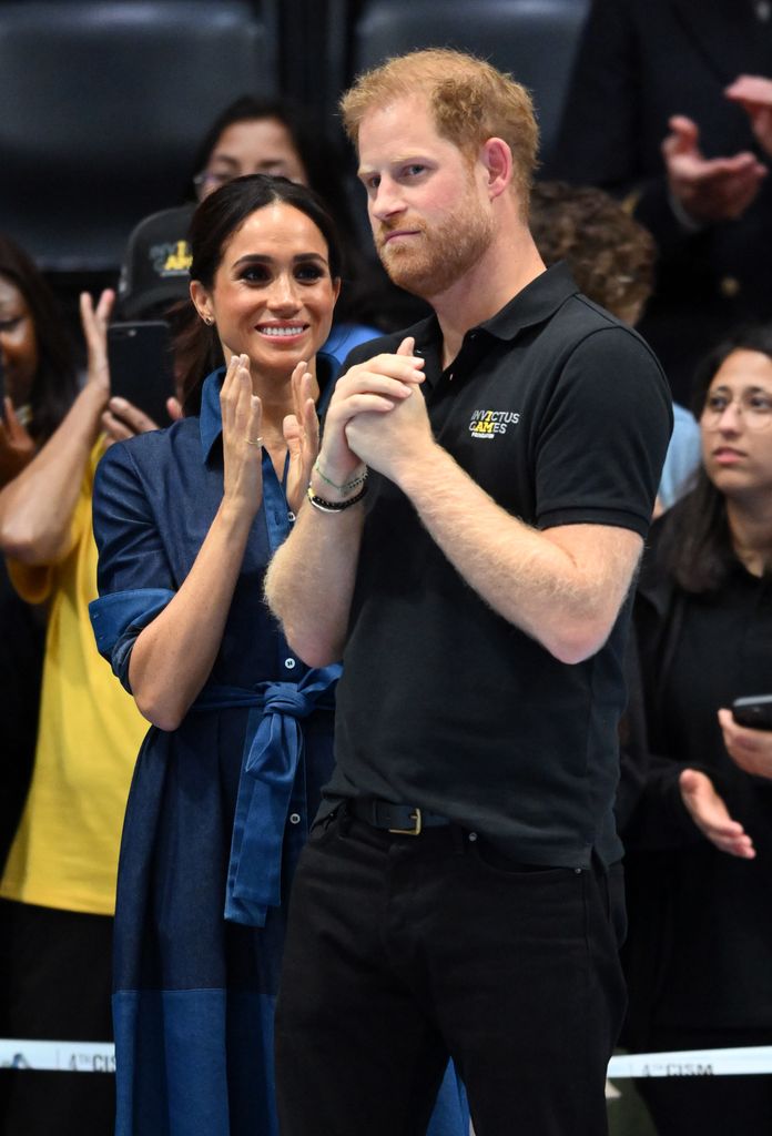 Meghan Markle clapping next to a grumpy(ish) Prince Harry