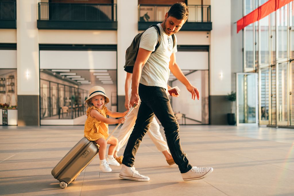 Un père fait rouler une valise avec une petite fille assise dessus à travers un aéroport