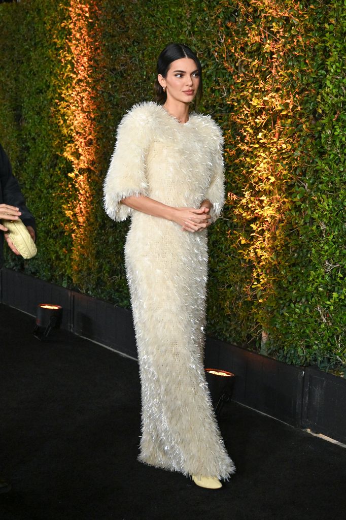 Kendall Jenner wearing Bottega Veneta walks down the red carpet. Her cream dress is textured with elbow length sleeves and she wears her hair in a slick back do. 