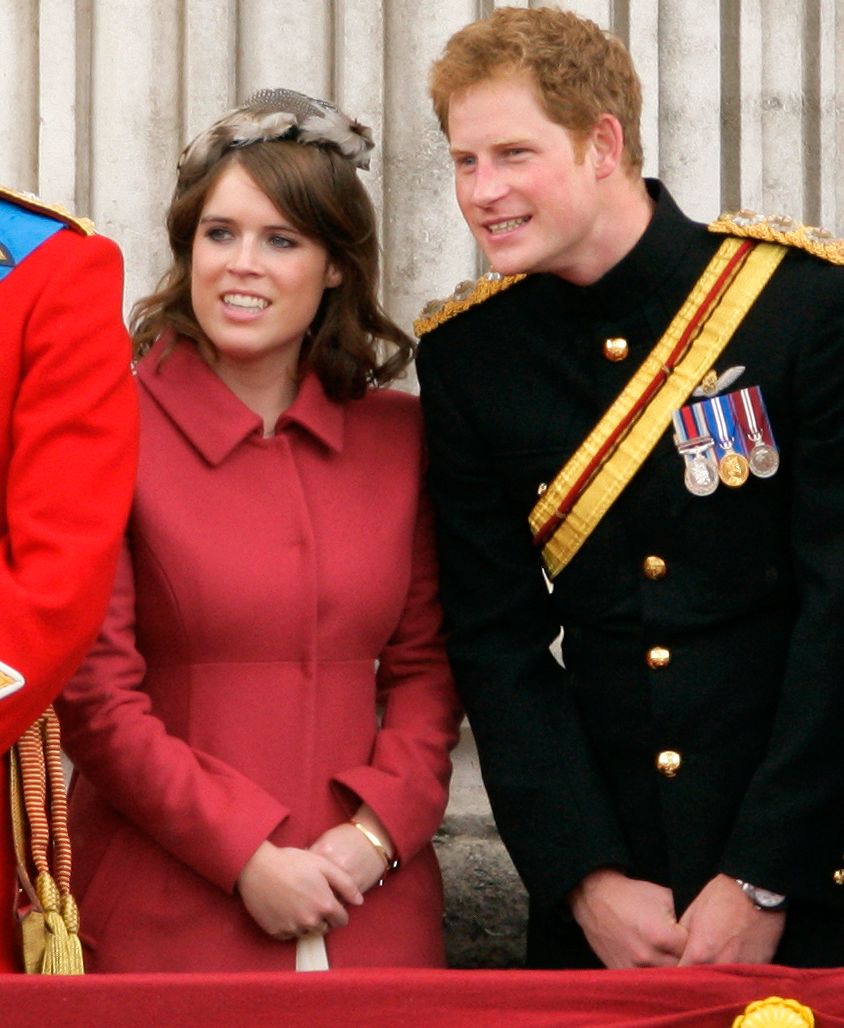 Princess Eugenie in a red jacket and Prince Harry in his black uniform stand on the balcony of Buckingham Palace