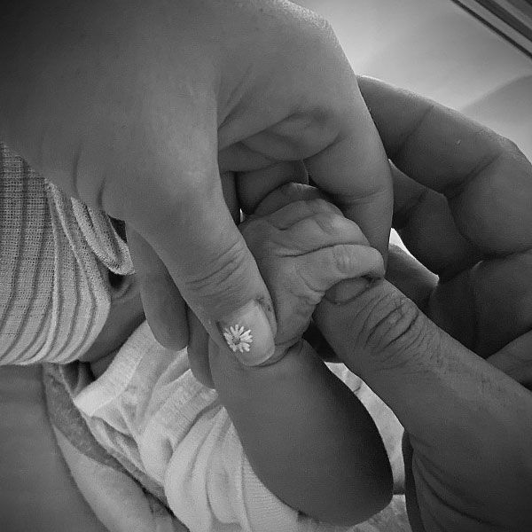 A black and white photo of two hands holding a little babys hand. One of the adults thumbs has a daisy painted on it