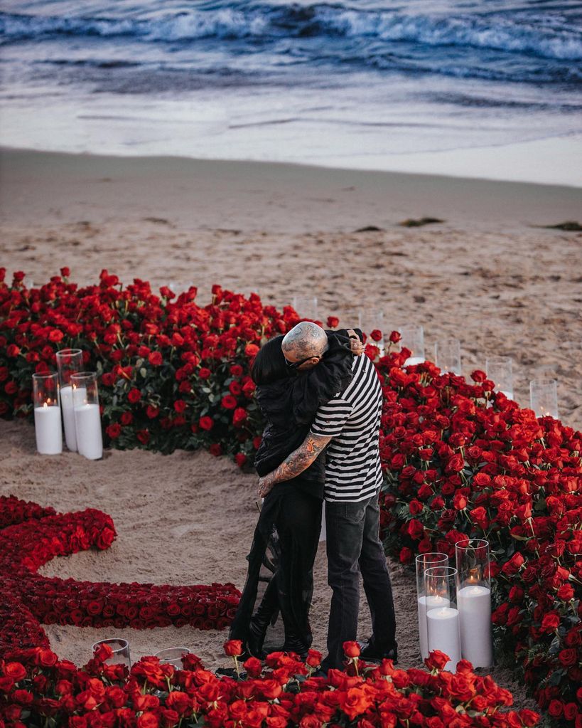 Travis Barker and Kourtney Kardashian hugging on the beach surrounded by roses
