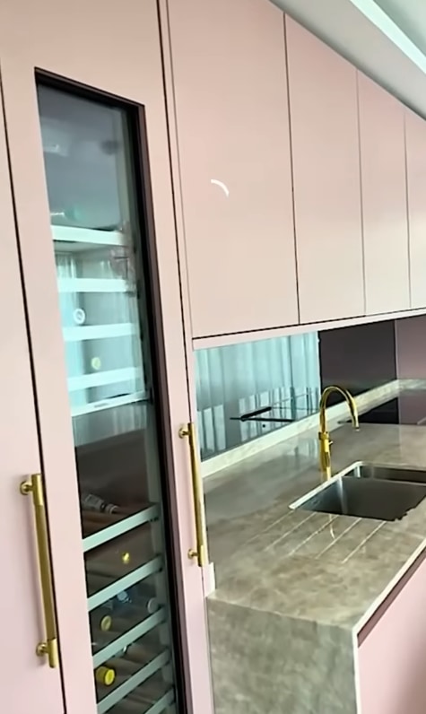 A modern kitchen with pale pink cabinets, gold handles, a built-in glass-front wine fridge, marble countertops, an undermount sink, and a gold faucet