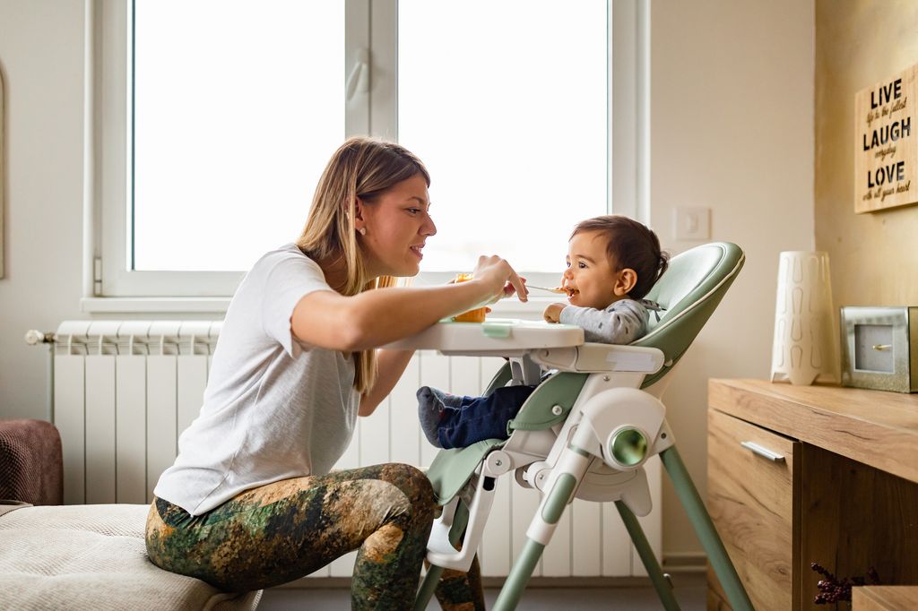 Family life. Young mother spoon feeding baby who is seated in a high chair in the living room
