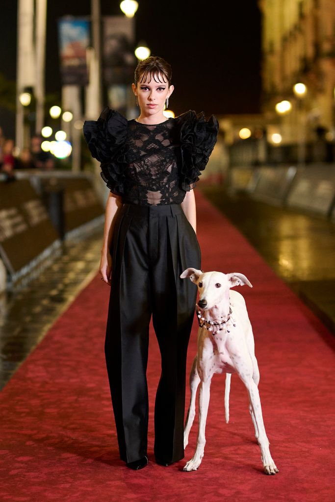 Actress Alba Planas wears  ensemble featured a high-fashion black lace top with voluminous ruffled sleeves, balanced by tailored black trousers. The structured yet soft textures created a dynamic contrast, making the look both playful and serious. The unexpected addition of a stylish greyhound companion made this moment even more memorable.