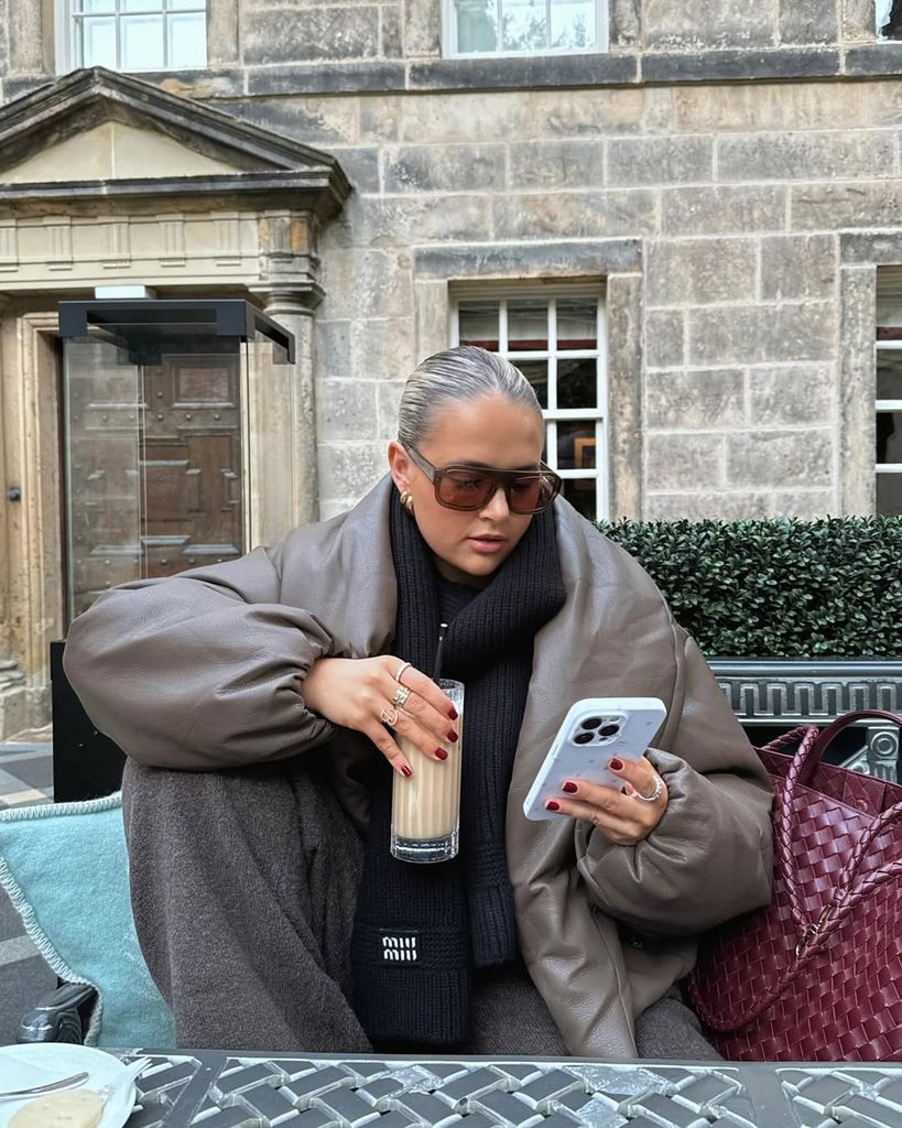 Molly-Mae sips a latte while scrolling on her phone. She’s wrapped in a brown bomber jacket, with slicked-back hair and a woven burgundy bag, exuding effortless sophistication.