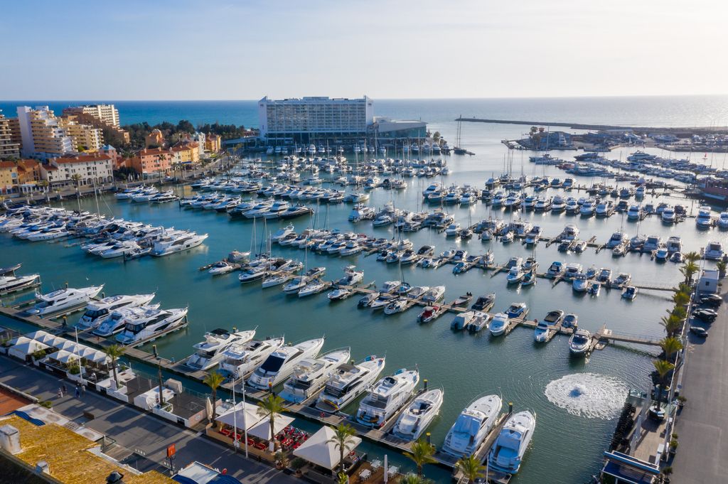 Aerial view of Vilamoura Marina in Portugal, showcasing luxury yachts docked along the waterfront with modern buildings and the Atlantic Ocean in the background.