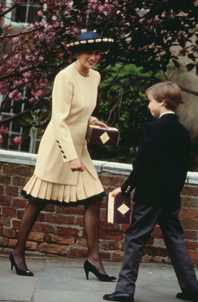 Princess Diana smiles at her son Prince William while wearing a butter yellow pleated suit with black trim, black pumps, sheer tights, and a wide-brimmed navy hat adorned with yellow ribbon.