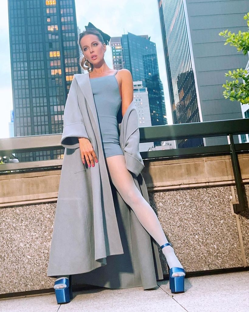 Kate Beckinsale wearing long blue coat and bodycon romper to pose against cityscape