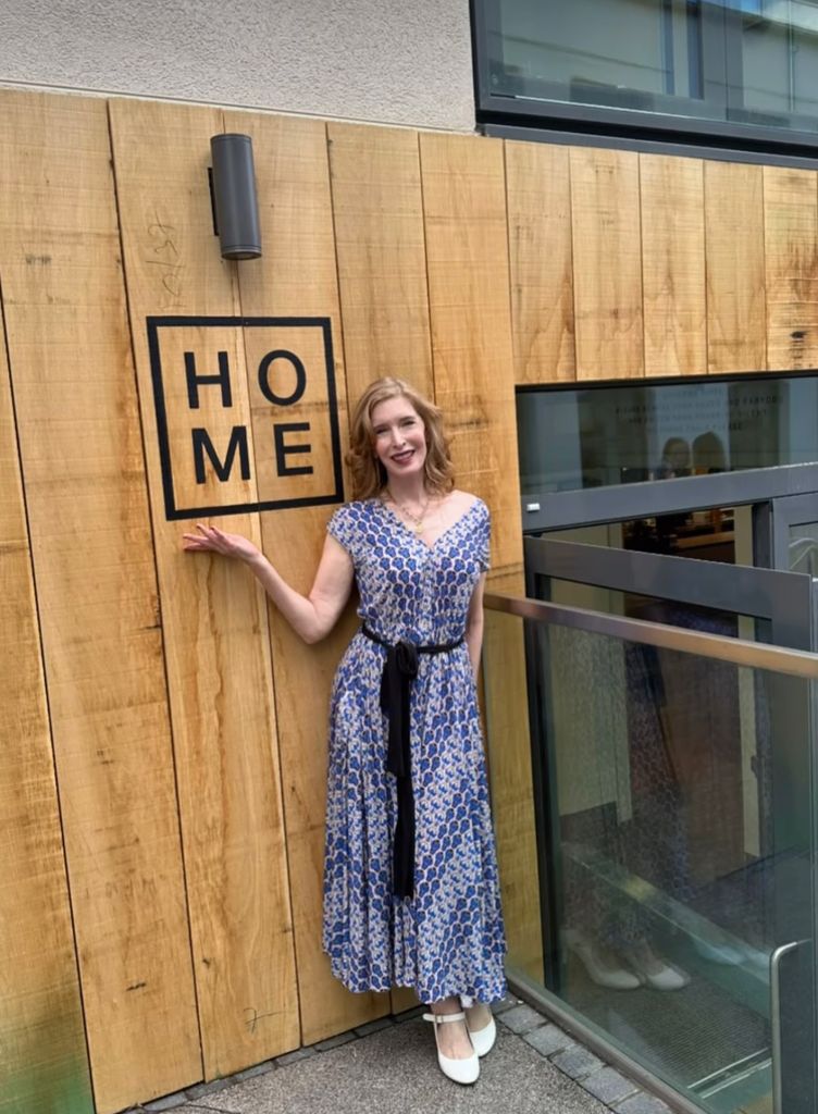 Woman in a printed jumpsuit next to a sign that says home 