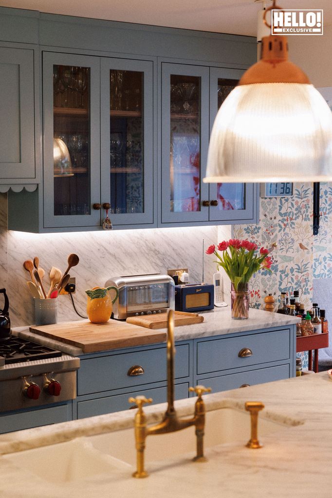 Nina Litchfield's kitchen at Notting Hill home. A stylish, traditional kitchen with blue cabinets, marble countertops, and gold hardware. There are flowers on the counter and a copper light fixture above.