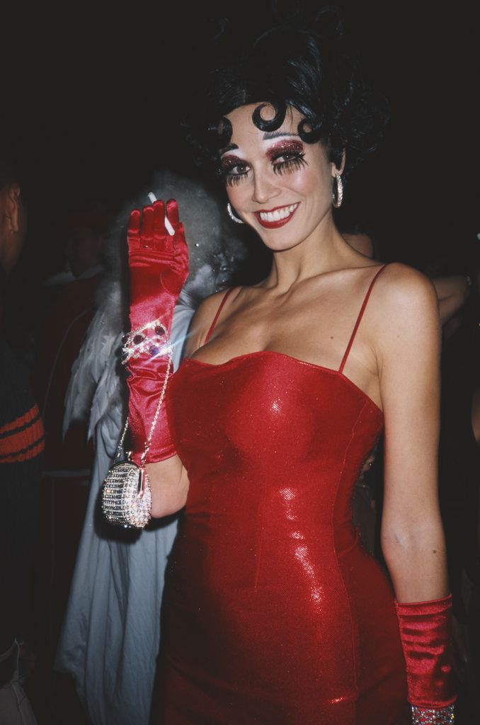 Heidi Klum wears a red, sparkly, fitted dress with curled black hair styled like Betty Boop. They accessorize with long red gloves and a small rhinestone purse, looking playful and glamorous.