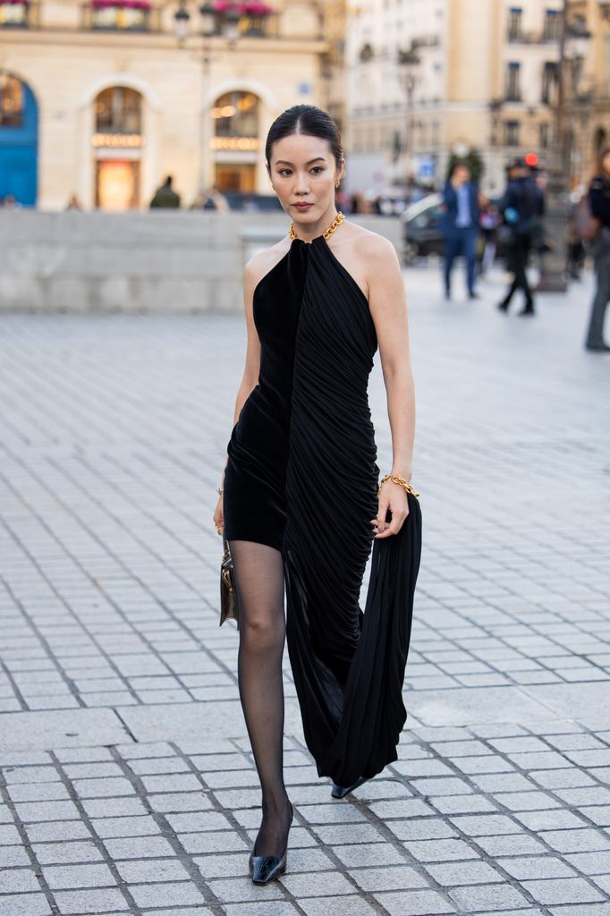 Jenny Tsang wears black dress with slit, golden bag outside Schiaparelli during Womenswear Spring/Summer 2025 as part of Paris Fashion Week