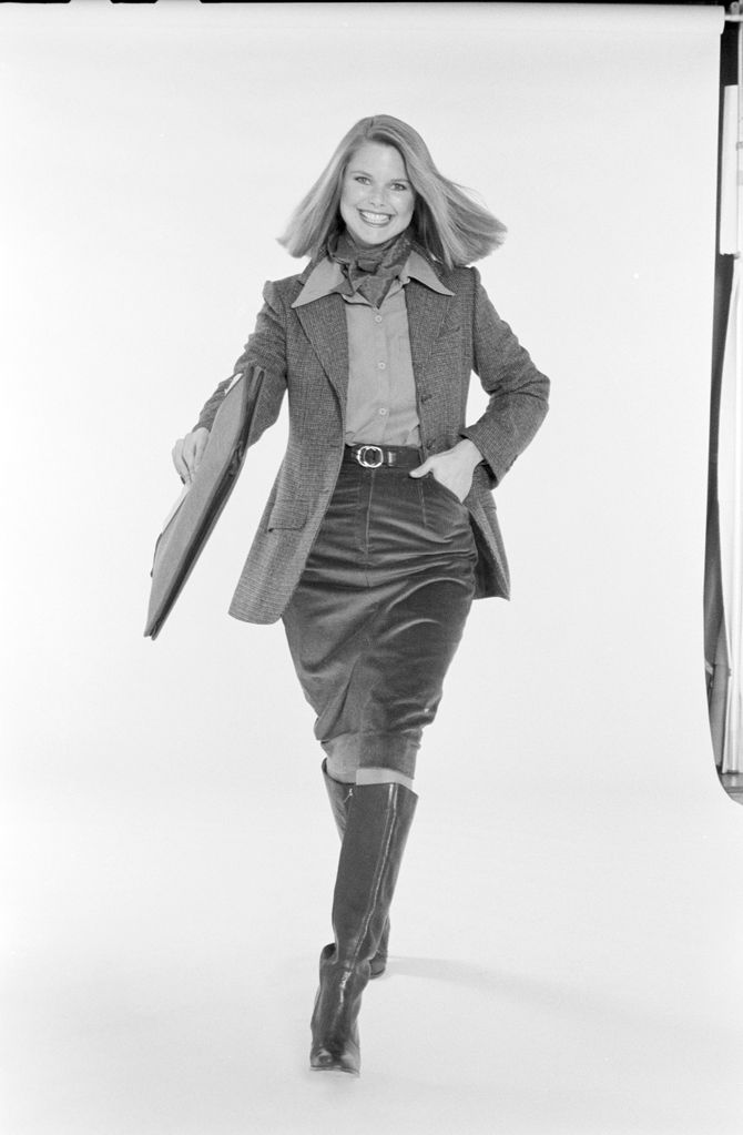 Christie Brinkley poses, dressed in business attire, against a white background, 1976. She wears a neckerchief, a tweed jacket over a button-down blouse, a corduroy skirt, and boots