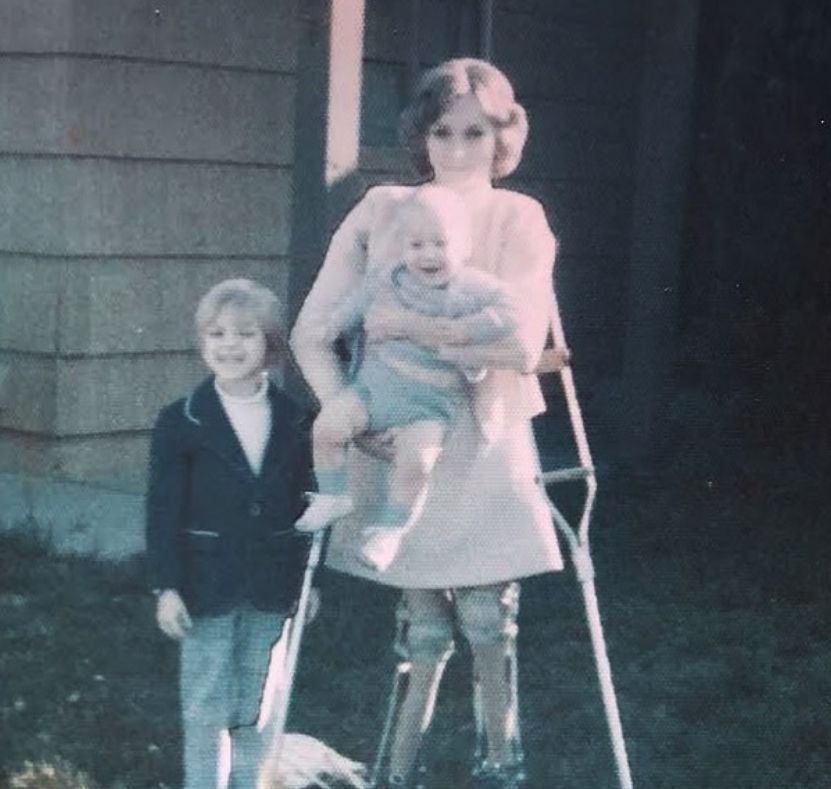 A woman on crutches carrying a baby while standing with a young boy