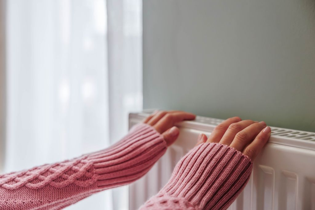 An unrecognisable woman in a pink warm jumper warming her hands at the heating radiator. Energy crisis and cold weather concept