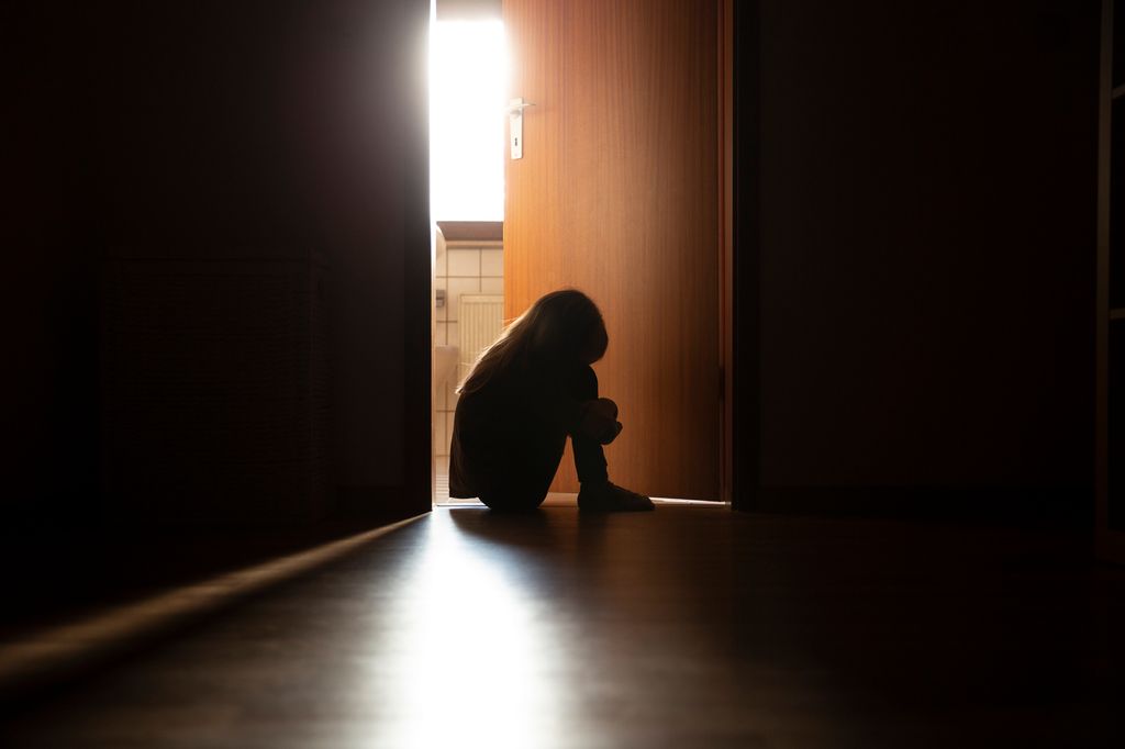 Despairing child sitting with head on knees in the dark frame of a doorway, backlit 