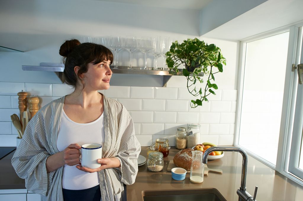 A woman smiles whilst holding a hot drink and looking out of the window of a zero waste kitchen.