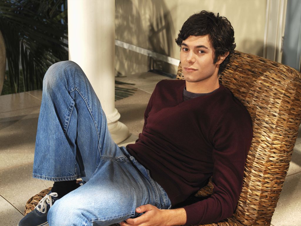 Adam Brody sitting on a wicker chair in a burgundy jumper