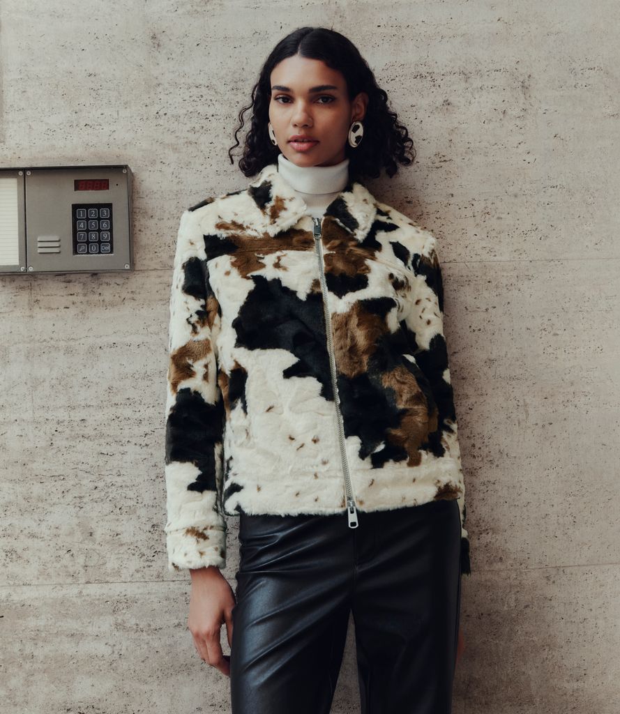 A model poses by a wall wearing a white polo neck, cow print jacket and black leather trousers. 