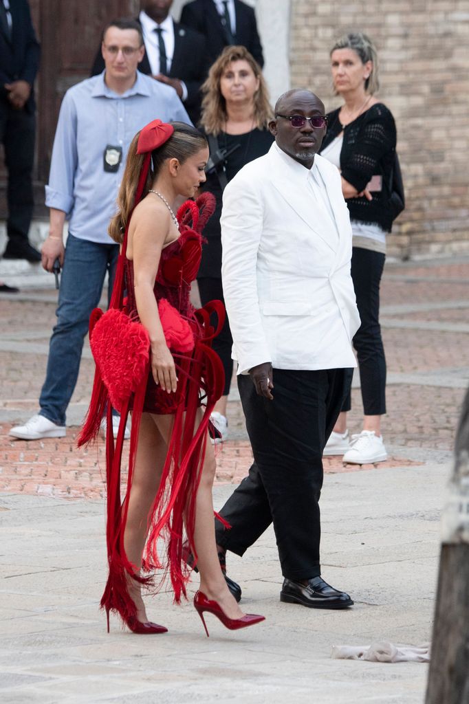 Edward Enninful arrives in a white jacket with Natasha Poonawalla wearing a bold red mini dress with heart-shaped details, fringe embellishments, and matching heels.