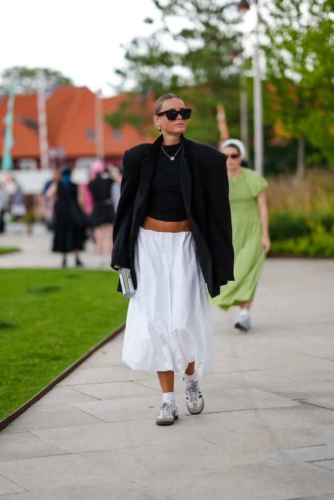 A guest wears black sunglasses, gold earrings, gold necklace, black shirt, black oversized blazer jacket, white midi skirt, white socks, white black striped Adidas Originals Sambas, outside Marimekko, during the Copenhagen Fashion Week Spring/Summer 2024-2025 on August 8, 2024 in Copenhagen, Denmark. (Photo by Edward Berthelot/Getty Images)