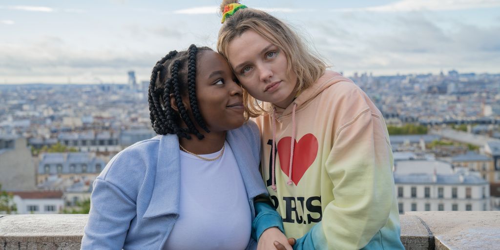 Tara and Darcy leaning on each other with an impressive city vista behind them, tara is smiling and looking at darcy, who is looking away thoughtfully