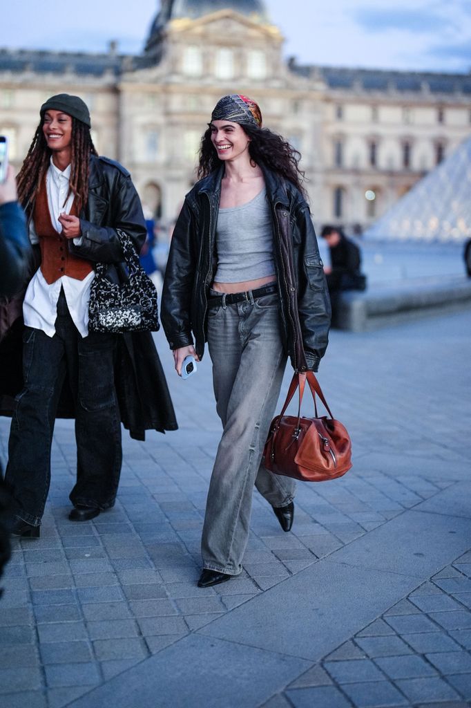  A model wears dark gray pattern head scarf, day gray tank top, shiny black oversized leather jacket, black leather belt, washed dark gray denim jean pants, shiny black pointed toe leather heels. shiny dark orange red leather handbag , outside Louis Vuitton, during the Paris Fashion Week Spring/Summer 2025 on October 1, 2024 in Paris, France (Photo by Edward Berthelot/Getty Images)