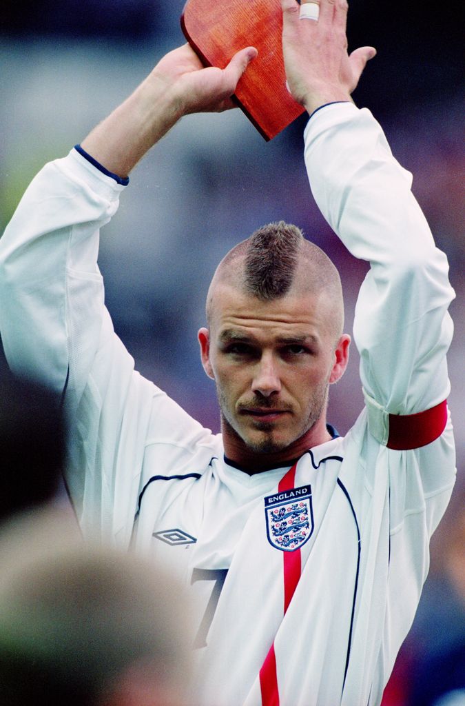 David Beckham raises his hands above his head in his football kit. His head is shaved into a faux mohawk. 